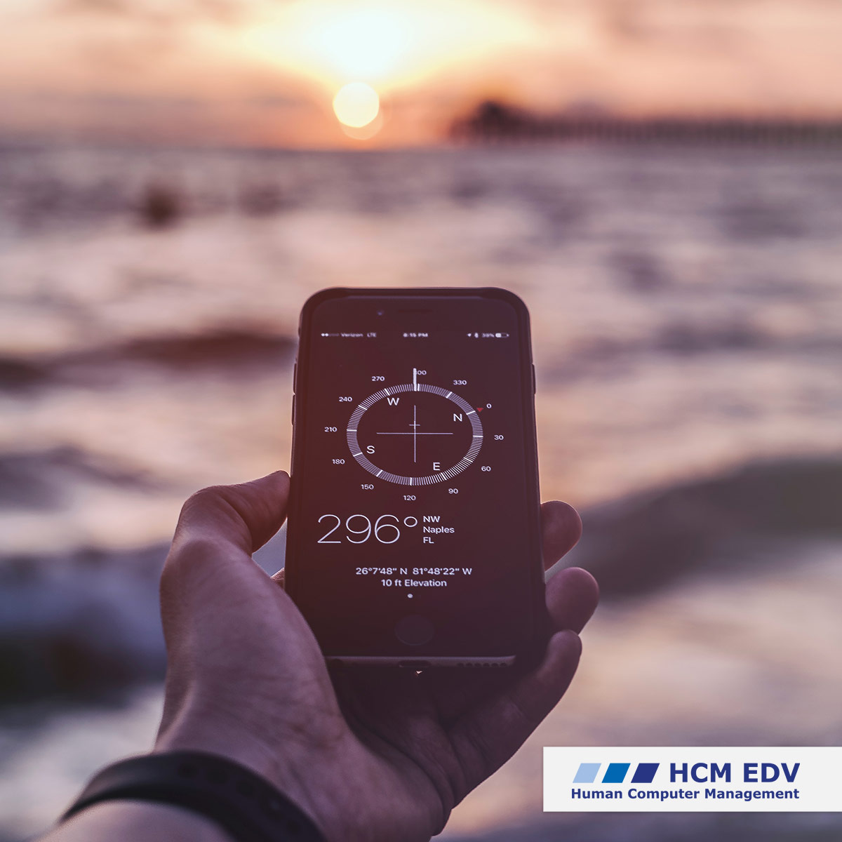 Eine Person hält ein Smartphone mit einer Kompass-App in Richtung der über dem Meer untergehenden Sonne