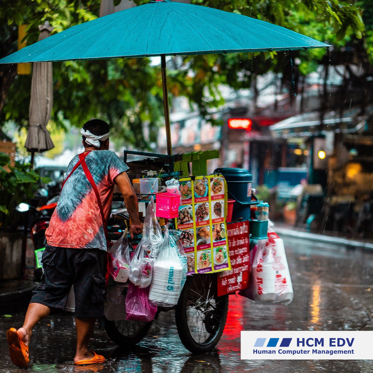 Asiatischer Mann schiebt seinen Essens-Wagen durch den Regen. Diesen ist durch einen großen türkisen Regenschirm geschützt.